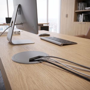 Pode incluir: Um passa-fios circular cinza-claro está embutido na superfície de uma mesa de madeira de carvalho claro. Vários cabos elétricos cinzas e pretos passam pela abertura. Um monitor de computador, teclado e mouse estão visíveis na mesa.