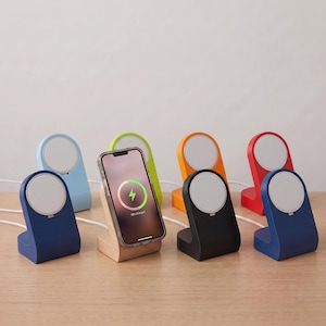 May include: A collection of seven wireless charging stands in various colors including light blue, lime green, orange, red, black, and dark blue, with one light wood stand holding a smartphone displaying a green circular charging icon.