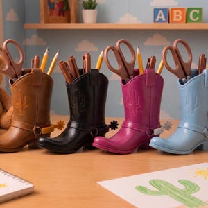 Puede incluir: Cuatro portalápices con forma de bota de vaquero en marrón, negro, magenta y azul claro. Cada bota presenta un diseño de cactus en relieve en el lateral, una espuela decorativa en el talón y contiene tijeras y lápices. Están en fila.