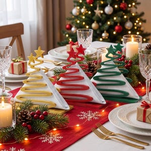 May include: Three triangular napkin holders shaped like Christmas trees in gold, red, and green. Each holder features a star at the top and a spiral wire design that holds white napkins, arranged on a red table runner with holiday decor.