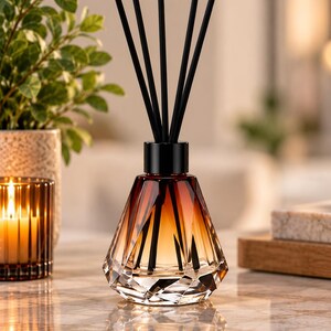 May include: A glass reed diffuser with a faceted, diamond-like shape featuring a gradient color transition from clear at the base to a deep amber at the top. It includes a black collar and five slender black reeds extending from the opening.