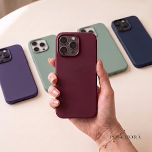 Peut inclure: Une collection de cinq coques de smartphone unies dans des tons violet profond, vert sauge, bordeaux et bleu marine. La coque bordeaux est tenue au premier plan, montrant une découpe pour triple objectif et une finition mate lisse.