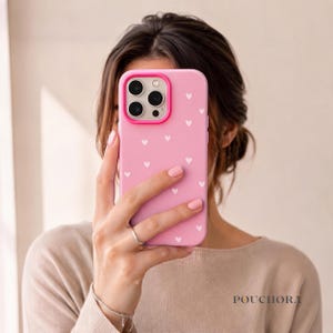 Pode incluir: Uma capa de smartphone rosa claro com um padrão de pequenos corações brancos e uma moldura de câmara rosa brilhante. A capa tem uma abertura para três lentes de câmara. A palavra POUCHORA está impressa em letras pretas no canto inferior.