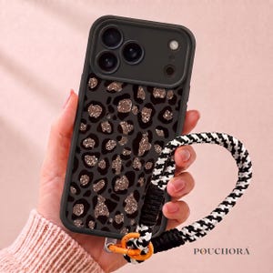Op de afbeelding: Een zwarte smartphonehoes met een donkergrijs luipaardpatroon en glinsterende roségouden glitteraccenten. De hoes bevat een zwarte camerabehuizing en een afneembare polsband met een zwart-wit gevlochten ontwerp en een oranje clip.