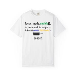 以下が含まれることがあります： 黒、緑、青のプログラミングコードテキスト「focus_mode.enable(); // deep work in progress System.out.println("100%Loader");」がプリントされた白い半袖Tシャツ。その下には100%の黒いバッテリーアイコンと「Loaded」という文字が配置されています。