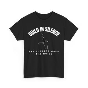 Può includere: Una t-shirt nera a maniche corte con testo bianco che recita BUILD IN SILENCE e LET SUCCESS MAKE THE NOISE, con una grafica a linee bianche di una silhouette di un volto con un dito davanti alle labbra in un gesto di silenzio.