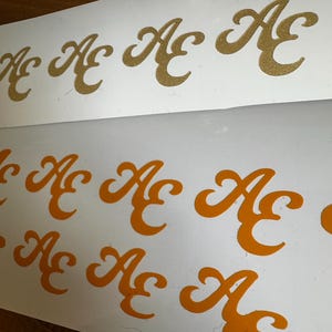 Könnte beinhalten: Ein Set aus Aufklebern auf weißen Trägerfolien, die die Buchstaben AE in einer stilisierten Schreibschrift zeigen. Die oberste Reihe besteht aus goldglitzernden Buchstaben, während die unteren beiden Reihen einfarbige orangefarbene Buchstaben in derselben dekorativen Schriftart aufweisen.