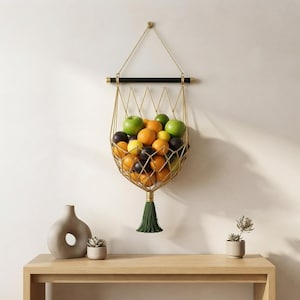 Puede incluir: Una cesta de frutas de metal dorado montada en la pared con un diseño de alambre geométrico, suspendida por una cadena dorada de una barra horizontal negra. La cesta contiene naranjas frescas, manzanas verdes y ciruelas oscuras, con borla verde.