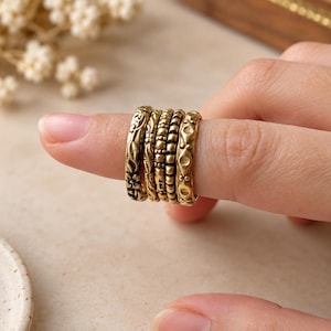 May include: A set of four stacked rings in a muted gold tone. Each ring features a distinct texture: one with a floral pattern, one with a leaf motif, one with a beaded design, and one with a repeating diamond shape with small circular accents.