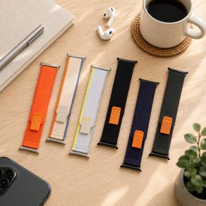 Può includere: Sei cinturini di ricambio per smartwatch, disposti in fila. I cinturini presentano una trama in tessuto intrecciato nei colori arancione, beige, grigio chiaro, nero, blu scuro e nero, ciascuno dotato di una linguetta arancione e connettori.