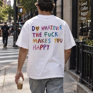 Pode incluir: Uma t-shirt branca de manga curta com o texto multicolorido DO WHATEVER THE FUCK MAKES YOU HAPPY nas costas. As letras são apresentadas num estilo desenhado à mão com dois pequenos rostos em forma de coração integrados no design.