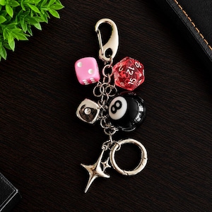 Puede incluir: Un llavero de metal plateado con varios dijes que incluyen un dado rosa de seis caras, un dado rojo translúcido de veinte caras, una bola de billar número ocho, un cubo metálico con una piedra transparente y una estrella de cuatro puntas.