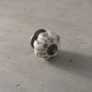 May include: A pumpkin-shaped cabinet knob featuring a white ceramic surface with a dark brown crackle pattern. The knob includes a dark metallic base and a small, circular, ribbed metal accent piece centered on the front of the ceramic body.
