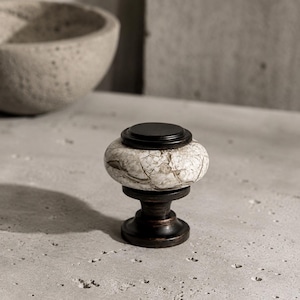 May include: A cabinet knob featuring a round, textured white and grey stone center with a cracked pattern. The knob has a dark brown, metallic base and a matching dark brown, flat circular top piece, creating a contrast between the stone and metal.