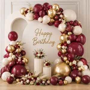 May include: A balloon arch decoration featuring burgundy, cream, and metallic gold balloons accented with floral arrangements. A white circular backdrop displays the gold script text Happy Birthday. Two fluted pedestals hold floral centerpieces.