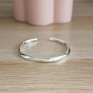 Op de afbeelding: Een zilverkleurige open armband met een gladde, gepolijste afwerking en een subtiele, golvende vorm. De band heeft een doorlopend, vloeiend ontwerp dat zachtjes buigt, wat zorgt voor een eenvoudig en reflecterend metalen oppervlak.