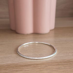 Puede incluir: Una pulsera tipo brazalete circular y delgada de plata con una superficie texturizada y brillante. El brazalete presenta un diseño de aro simple y continuo con un brillo metálico, reflejando la luz en su textura exterior granulada.