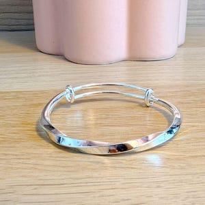 May include: A silver-toned expandable bangle bracelet featuring a polished, twisted metal design. The bracelet has two circular loops at the top where the metal bands overlap, allowing the diameter to be adjusted for a comfortable fit on the wrist.