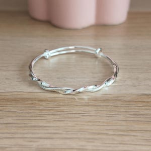 Puede incluir: Una pulsera tipo brazalete de color plateado con un diseño trenzado similar a una cuerda. La pulsera es circular y cuenta con dos pequeños remates redondeados donde se unen los extremos de la banda metálica, creando una superficie pulida.