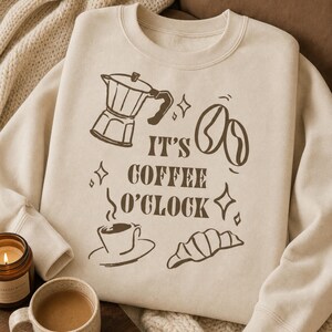 Puede incluir: Una sudadera de cuello redondo en color beige con un estampado gráfico en marrón oscuro que muestra una cafetera italiana, granos de café, una taza de café humeante y un cruasán. El texto IT'S COFFEE O'CLOCK está centrado y rodeado de estrellas.