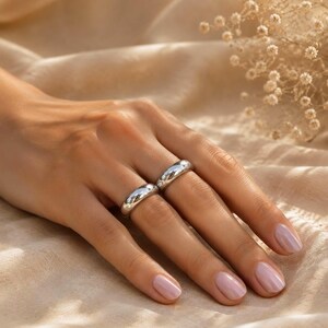 May include: Two polished silver-tone dome rings with a smooth, rounded surface. The rings feature a thick, solid band design with a high-shine metallic finish, worn on the middle and ring fingers, showcasing a simple, reflective, and curved aesthetic.