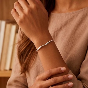 Può includere: Un bracciale rigido in argento lucido caratterizzato da un design continuo a onde morbide. Il metallo presenta una superficie liscia e riflettente che cattura la luce, creando sottili riflessi lungo la forma curva e sinuosa dell'anello.