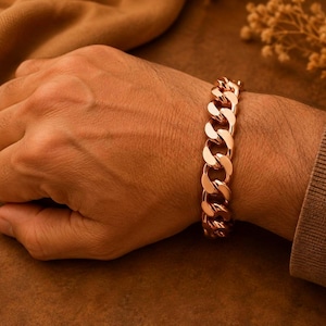 May include: A polished rose gold-tone curb chain bracelet featuring thick, interlocking oval links. The bracelet has a high-shine metallic finish and is secured around the wrist, showcasing a classic, heavy-duty link design with smooth edges.