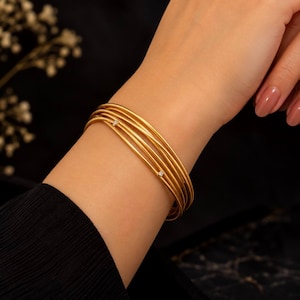 Può includere: Un bracciale rigido in oro a più fili composto da cinque cerchi sottili, lisci e circolari che avvolgono il polso. Due di questi cerchi sono impreziositi da una piccola pietra preziosa trasparente dal taglio rotondo incastonata nell'oro.