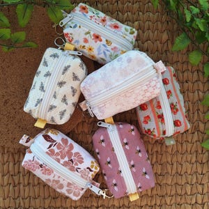 May include: A collection of six small rectangular fabric pouches with white zippers and metal clips. The pouches feature various patterns including floral prints in pink, orange, and green, a bee print on cream fabric, and a quilted pink bee print.
