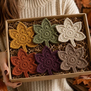 May include: A set of six crochet leaf coasters in a cardboard box filled with brown paper filler. The coasters are shaped like maple leaves and come in mustard yellow, olive green, cream, burnt orange, deep purple, and brown yarn colors.