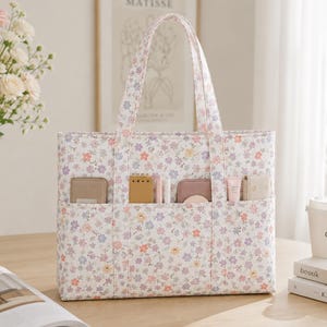 May include: A rectangular tote bag featuring a white base with a small-scale floral pattern in shades of pink, purple, and orange. The bag has two shoulder straps and three exterior front pockets containing various small personal items.