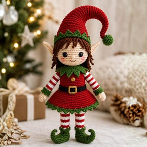 Puede incluir: Una muñeca elfo de ganchillo con un gorro rojo puntiagudo con un pompón verde, pelo castaño y grandes ojos negros. La muñeca lleva un vestido rojo con cuello y dobladillo verdes, un cinturón negro con hebilla amarilla y mangas y mallas a rayas rojas y blancas.