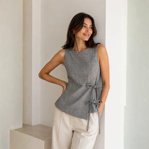 Puede incluir: Una blusa sin mangas con estampado de cuadros glen en blanco y negro, cuello redondo y cierre lateral estilo cruzado asegurado con dos juegos de finas cintas de tela en el lado izquierdo, combinada con pantalones beige claro.