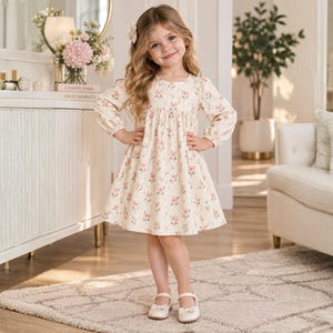 May include: A cream-colored long-sleeve dress featuring a repeating pattern of small pink flowers and green leaves. The dress has a round neckline, a gathered waistline, and a flared skirt. It is paired with light-colored flat shoes with a strap.