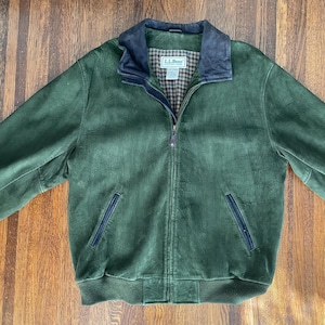 May include: A dark green suede jacket featuring a navy blue collar, a full-length front zipper, and two side pockets with navy blue trim. The interior lining displays a brown and tan plaid pattern with an L.L.Bean brand label visible.