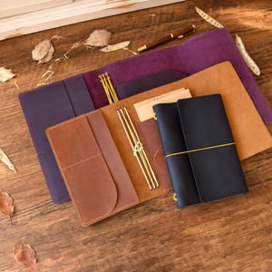 Puede incluir: Una colección de fundas de cuero para cuadernos en morado, tostado y azul oscuro, con bolsillos internos y encuadernaciones de cordón elástico amarillo. Una pluma estilográfica marrón descansa sobre la funda morada y un cuaderno azul oscuro está sujeto con una banda amarilla.