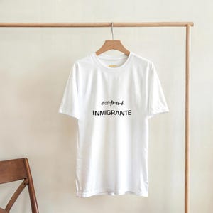 Può includere: Una t-shirt bianca a maniche corte appesa a una gruccia di legno. La parte anteriore della maglietta presenta la parola "expat" con una linea che la attraversa, posizionata sopra la parola "INMIGRANTE" stampata in grassetto, nero, maiuscolo.