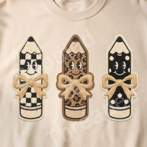 May include: A beige crewneck sweatshirt featuring a graphic of three anthropomorphic pencils. Each pencil has a smiling face and a tan bow. The left pencil has a black and white checkerboard pattern, the middle has a leopard print, and the right is black with white polka dots.