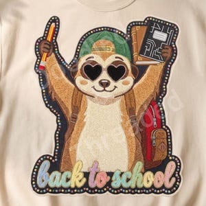 May include: A cream-colored crewneck sweatshirt featuring a graphic of a meerkat wearing a green cap and heart-shaped sunglasses. The meerkat holds a pencil and books, wears a backpack, and is surrounded by the text back to school in colorful letters.