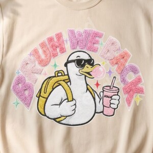Könnte beinhalten: Ein beigefarbenes Sweatshirt mit Rundhalsausschnitt und einer Grafik einer weißen Ente mit Sonnenbrille, gelbem Rucksack und einem rosa Getränk mit Kaugummiblase. Der Text BRUH WE BACK ist in einer strukturierten rosa und lila Schrift darüber gedruckt.
