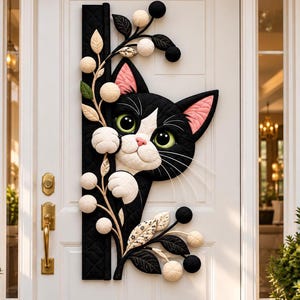 May include: A black and white cat wall hanging with green eyes and pink inner ears, peeking from behind a vertical black quilted panel decorated with cream-colored spherical blossoms and dark leaves, mounted on a white door.