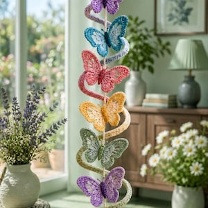 May include: A vertical hanging mobile featuring six crocheted butterflies in purple, blue, pink, yellow, green, and light purple. The butterflies are connected by a spiraling yarn strand and a central cord, finished with a clear crystal prism at the bottom.