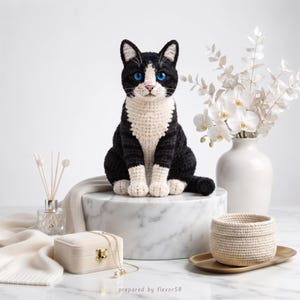 Puede incluir: Una figura de gato tejida a ganchillo a mano con hilo negro y blanco, orejas puntiagudas y ojos azul brillante. El gato está sentado en posición vertical con patas y pecho blancos, mostrando un tejido detallado en todo su cuerpo.