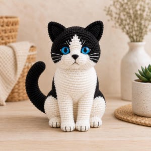 May include: A black and white crocheted cat figurine with bright blue eyes. The cat is sitting in an upright position with its tail curved upwards. It features a white chest, face, and paws, with black ears, head, back, and tail. The yarn texture is visible.