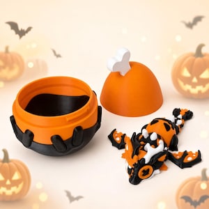 Puede incluir: Un recipiente naranja y negro con forma de caldero con una tapa a juego rematada con un hueso blanco, acompañado de una pequeña figura de dragón articulada con segmentos naranjas, negros y blancos con estructuras detalladas de alas y cola.
