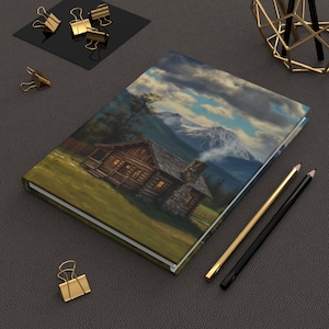 May include: A rectangular hardcover journal featuring a printed landscape illustration of a rustic log cabin with glowing windows, a stone chimney, and snow-capped mountains under a cloudy sky, accompanied by two pencils and gold binder clips.