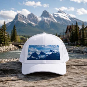 May include: A white trucker hat featuring a rectangular graphic print of snow-capped mountains and the text Canmore, Alberta. The cap has a solid front panel, a curved brim, and a mesh back panel, providing a structured look and breathable design.