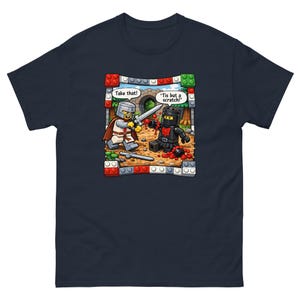 May include: A navy blue t-shirt featuring a graphic of two toy knights in a duel. One knight in silver armour says 'Take that!' while the other in black armour says 'Tis but a scratch!' The scene is framed by colourful plastic building blocks.