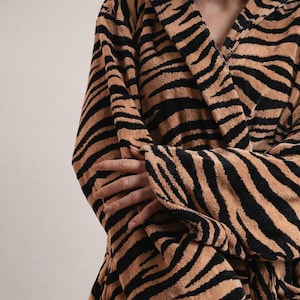 May include: A plush bathrobe featuring a bold tiger stripe pattern in shades of black and tan. The garment has long sleeves, a wide collar, and a textured fabric surface that covers the torso and arms in a repeating animal print design.