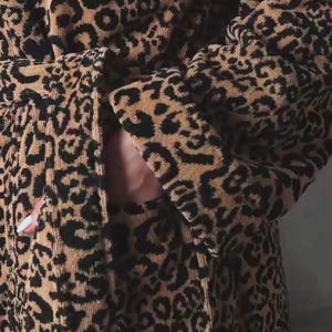 May include: A plush bathrobe featuring a prominent black and tan leopard print pattern. The garment includes long sleeves with wide cuffs, a matching tie belt at the waist, and a visible side pocket integrated into the textured fabric design.